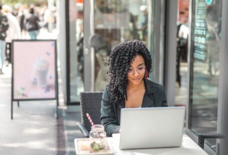 High-End Shopping - High angle of pensive African American female freelancer in glasses and casual clothes focusing on screen and interacting with netbook while sitting at table with glass of yummy drink on cafe terrace in sunny day