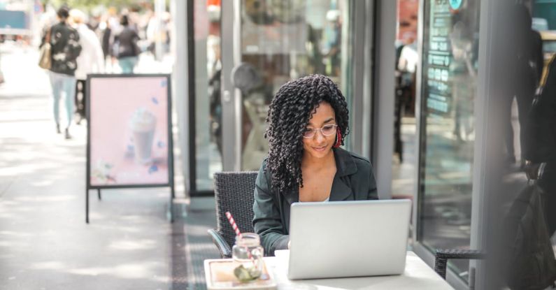 High-End Shopping - High angle of pensive African American female freelancer in glasses and casual clothes focusing on screen and interacting with netbook while sitting at table with glass of yummy drink on cafe terrace in sunny day